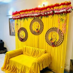 For a home mehndi or haldi function, I can create a vibrant photo corner like this one. This setup includes a bright yellow backdrop with matching sofa covers, decorative dreamcatcher-style hangings, and a floral border on top.