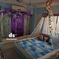 A cozy and romantic room setup with a beautiful canopy tent over the bed, decorated with fairy lights and ivy. A purple and silver balloon and fringe backdrop adds a celebratory touch on the opposite wall.