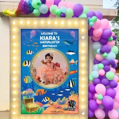 A personalized welcome board for Kiara's underwater-themed party. The board features a photo of the birthday girl and is framed with lights and a balloon garland, making it a great photo spot for guests.