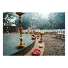 The pathway of a wedding ceremony is lined with tall brass lamps adorned with flowers. The warm glow of the lamps and the floating petals in the urlis create a magical and sacred walkway.