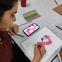A student uses a reference on her phone to carefully draw a flamingo, focusing on capturing the bird's elegant curves with colored pencils.