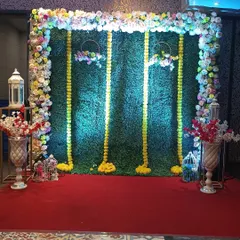 A Haldi backdrop combining a faux green wall with marigold garlands and a floral frame. Decorative lanterns and birdcages on the side add extra detail to the setup.