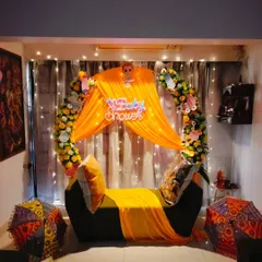 A vibrant home baby shower decoration, featuring a sofa decorated with bright yellow drapes, a floral arch, and a "Baby Shower" neon sign.