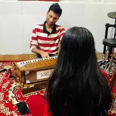 A coach provides individual attention to a vocal student, guiding her through exercises on the harmonium. This personalized feedback is key to vocal development.
