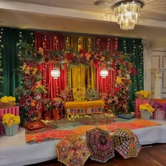 A vibrant stage designed for a Mehendi function. The decor is built around a rich palette of red, orange, and green, with a dense floral arch and colourful Rajasthani umbrellas adding a traditional, celebratory touch.