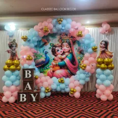 A traditional Radha Krishna theme for a baby shower. This setup includes a custom printed backdrop, character cutouts, and a circular balloon arch in soft pink and blue, accented with gold balloons and wooden 'BABY' blocks.