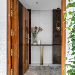 The entrance to Dr. Harsha's home offers a glimpse of the minimalist luxury within. A custom carved wooden door opens to a foyer with clean lines, a sleek console table, and contrasting dark wood paneling.