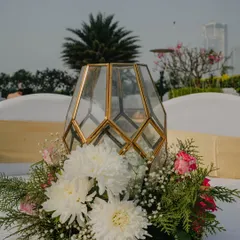A simple yet elegant centerpiece for an outdoor event. A geometric glass and gold lantern is surrounded by a wreath of fresh white chrysanthemums, pink roses, and greenery, set on a rustic wood slice base.