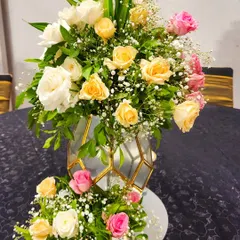 A beautiful floral arrangement in a geometric gold and glass vase. The bouquet features a soft palette of yellow, white, and pink roses, perfect for an elegant reception or gala dinner.