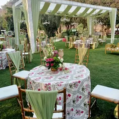 An elegant outdoor seating arrangement for a day wedding. The tables are covered with floral-printed cloths, and the chairs are decorated with sage green drapes.