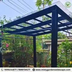 The compact sheet pergola is set within a lush garden, creating a perfect shaded retreat for enjoying the outdoors. The minimalist design blends seamlessly with the natural surroundings.