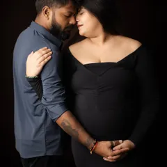 An intimate close-up of a couple in a dark studio setting. Dressed in dark, coordinating outfits, they share a tender forehead-to-forehead moment, their hands joined over the baby bump.