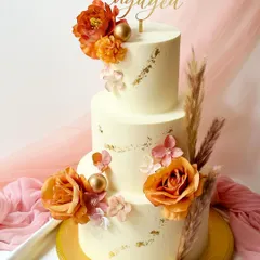 A magnificent three-tier engagement cake in off-white buttercream, decorated with gold leaf accents and a cascade of orange-toned roses and dried pampas grass.