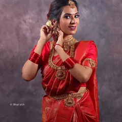 Capturing a candid moment during a traditional bridal shoot. The model adjusts her jhumka earring, showcasing the detailed gold jewelry and the rich texture of the red silk saree.