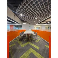 A long collaborative table set against a vibrant orange partition wall, designed for quick team huddles. The grid ceiling and patterned carpet define this zone within the larger workspace, demonstrating our use of design to guide function.