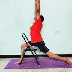 Veerabhadrasana 1 (Warrior 1) with the front thigh supported by a chair. This modification helps in squaring the hips and extending the spine upwards without straining the lower back.