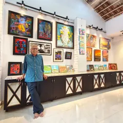 Senior cartoonist and art connoisseur Sridhar Comaravalli enjoying the "Vernissage Art Exhibition," leaning against a display of diverse artworks.