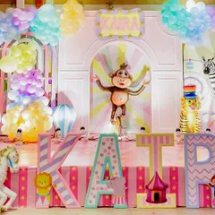A vibrant, pastel colored carnival stage with a monkey on a unicycle as the centerpiece. Large, custom letters spelling the birthday girl's name are decorated with different patterns and characters.