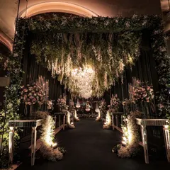 An absolutely magical entrance tunnel for a reception at Pullman New Delhi. The design features a ceiling of hanging florals and greenery, with a large chandelier and mirrored pedestals creating a forest of light.