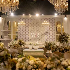 A view of the lounge area for the all-white wedding. We created a stunning backdrop with a custom geometric jaali pattern, framed by elegant white drapes and lit by chandeliers, with a classic white fountain adding a touch of serenity.