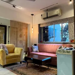 An eclectic living room with a mix of furniture styles, including a pink mid-century modern sofa and a yellow armchair. The lighting combines track lights with a decorative pendant light.