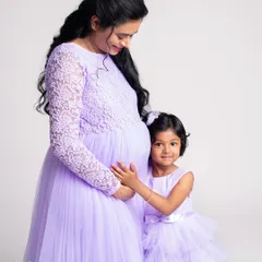 A mother and daughter share a special moment, both dressed in matching lavender gowns. The daughter gently touches her mother's baby bump, excited to become a big sister.