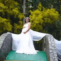 An angelic look for a mom-to-be, featuring a classic white maternity gown with a long, flowing train. This setup on a rustic bridge amidst lush greenery creates a serene and timeless portrait. The rental includes the gown and a matching floral hair accessory.