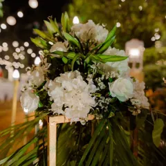 A beautiful floral arrangement of white lilies and hydrangeas on a gold stand, set against a backdrop of soft bokeh lights. This kind of detail adds a layer of sophistication and elegance to any event space.