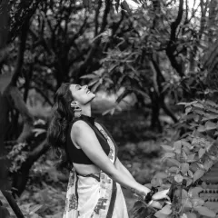 A dramatic black and white shot, capturing a free-spirited moment as the model looks up towards the sky in a lush, wooded area.