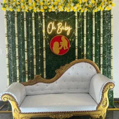 An elegant baby shower stage with a traditional touch. The setup features a beautiful white and gold chaise lounge set against a green foliage wall with an "Oh Baby" neon sign and floral garlands.