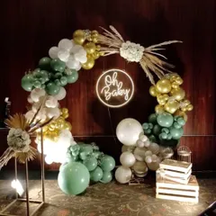 An elegant and modern baby shower backdrop, with a circular balloon arch in sage green, white, and gold, and a neon "Oh Baby" sign.