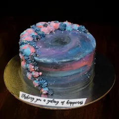 A galaxy-themed cake with a message "Wishing you a galaxy of happiness". The buttercream is painted in shades of blue, purple, and pink, with a swirl of piped stars on top.