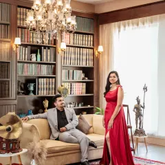 A sophisticated portrait in a classic library setting. The couple exudes elegance, with the bride in a striking red gown and the groom looking dapper, surrounded by books and a crystal chandelier.