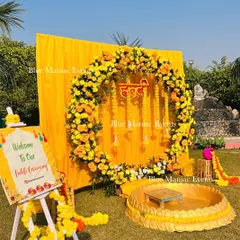 An outdoor Haldi ceremony setup featuring a circular floral wreath of marigolds against a bright yellow backdrop. The arrangement includes a personalized welcome sign and traditional seating for the rituals.