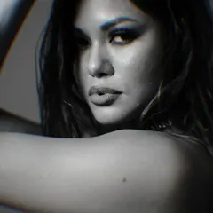 An intense, grainy black and white close-up portrait. The tight framing and dramatic shadows highlight the model's strong features and smoky eye makeup, creating a raw and emotional image.