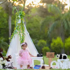 An outdoor first birthday setup with a teepee, books, and stuffed animals. The natural light and beautiful greenery create a magical setting.