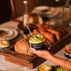 A jar of our Green Goddess Hummus, a vibrant and creamy dip held against a backdrop of a breakfast spread. It's a healthy and delicious addition to any meal.