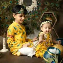 Two siblings in matching yellow Krishna-themed outfits interact playfully during their festive Janmashtami studio session.