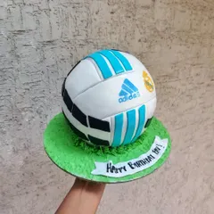 A fully spherical football cake, ready for a birthday celebration. The details are all hand-painted to create a realistic look, sitting on a field of green buttercream grass.
