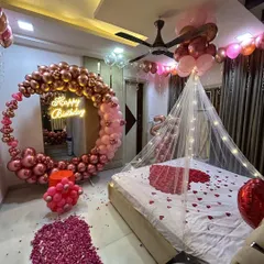 A wide view of a romantic birthday room setup. This decoration combines a large balloon ring, a bed canopy with lights, and extensive rose petal arrangements on the floor and bed for a grand romantic gesture.