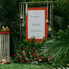 The entrance to a Telugu Nalugu ceremony, featuring a welcome board set amidst lush greenery and red roses. The hanging temple bells and traditional floral props add a divine and auspicious touch.