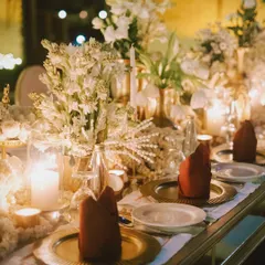 An elegant tablescape for a formal dinner, with gold charger plates, deep red napkins, and a centerpiece of white tuberose, all glowing by candlelight.