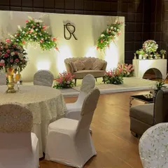 A full view of an elegant Roka stage setup. The design includes a clean white backdrop, asymmetrical floral arrangements, a custom couple's monogram, and a comfortable lounge sofa for the couple.