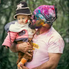 A side profile capturing the baby's curious expression as he looks at his father, whose face is covered in vibrant Holi colors. This image tells a story of a shared first experience.