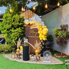 A sophisticated 40th birthday celebration in the garden. The setup includes a personalized backdrop, a black, gold, and silver balloon garland, and elegant decor pieces for a memorable milestone party.