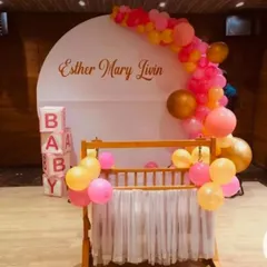 A modern and chic naming ceremony for "Esther Mary Livin". The clean white circular backdrop is accented with a colorful balloon garland, with a traditional wooden cradle at the forefront.