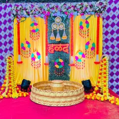 A bright and festive Haldi stage. The setup includes a yellow backdrop with a grass wall center, decorated with colorful hangings and marigold garlands.