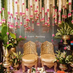 This is the complete setup for an intimate pre-wedding ceremony. It features ornate golden chairs, a canopy of floral strings, traditional brass lamps, and beautifully arranged offerings for the pooja.