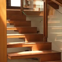 Sunlight streams across this teak wood staircase, highlighting the clean lines and warm grain of the material. The glass railing ensures safety without obstructing the view, maintaining the open and airy feel of the space.