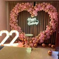 A chic birthday party decoration with a heart-shaped balloon arch in pink and rose gold. The setup is completed with a 'Happy Birthday' neon sign and large light-up numbers for the age.